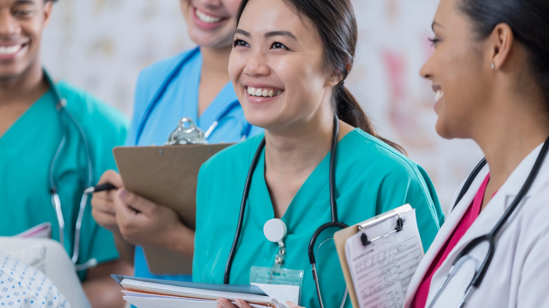 Group of smiling medical students and doctors in scrubs and white coats holding clipboards and stethoscopes, engaged in a positive discussion during clinical training