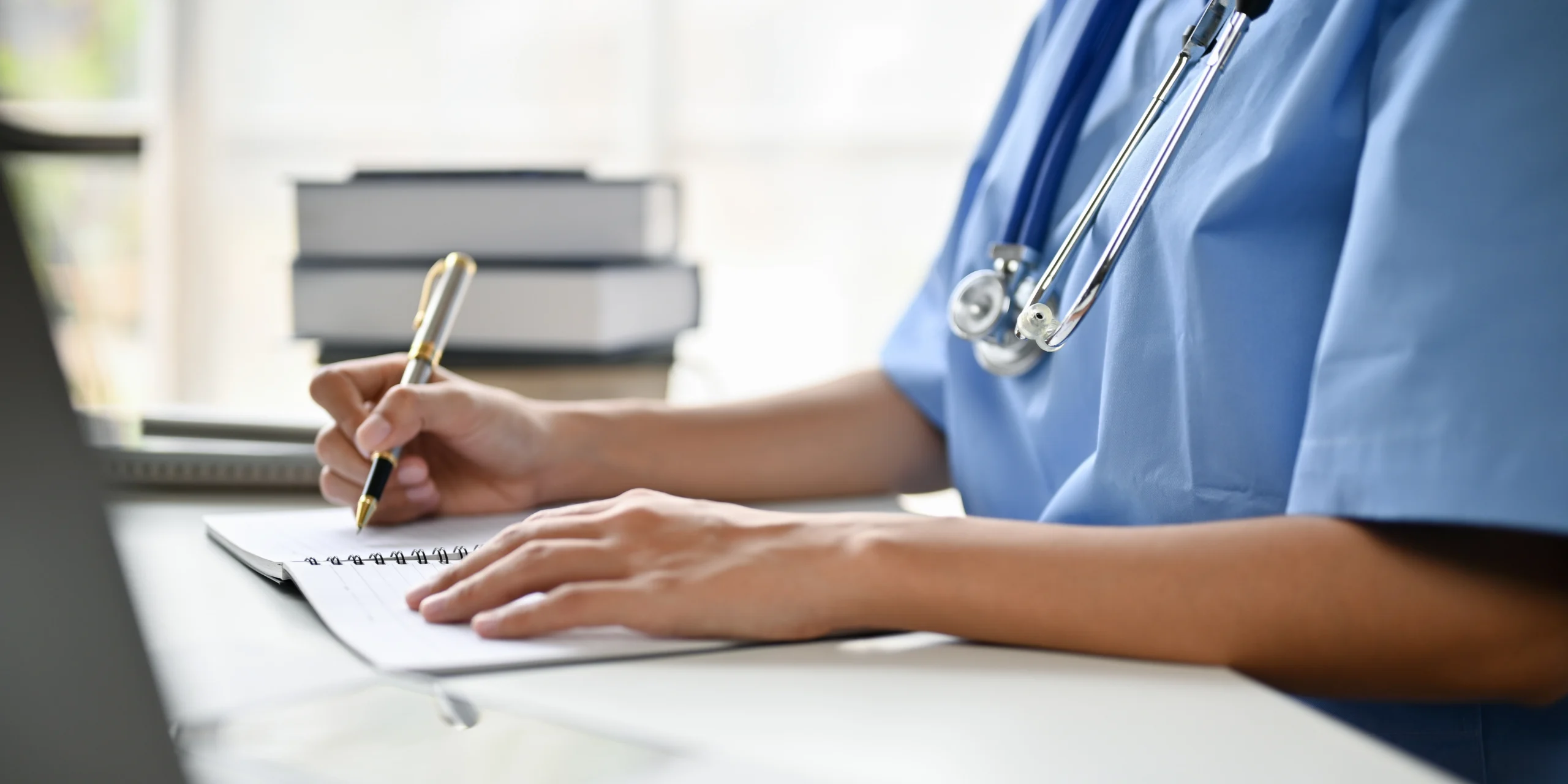 A healthcare professional in a blue scrubs and stethoscope writing notes on a spiral-bound notepad at a desk.