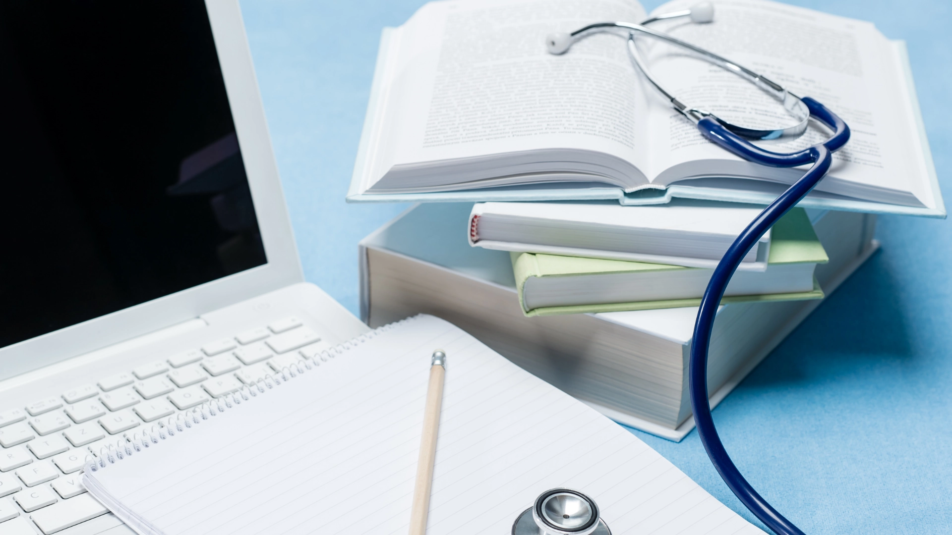 Medical study setup with a laptop, open notebook, pencil, stack of textbooks, and a stethoscope placed on an open book