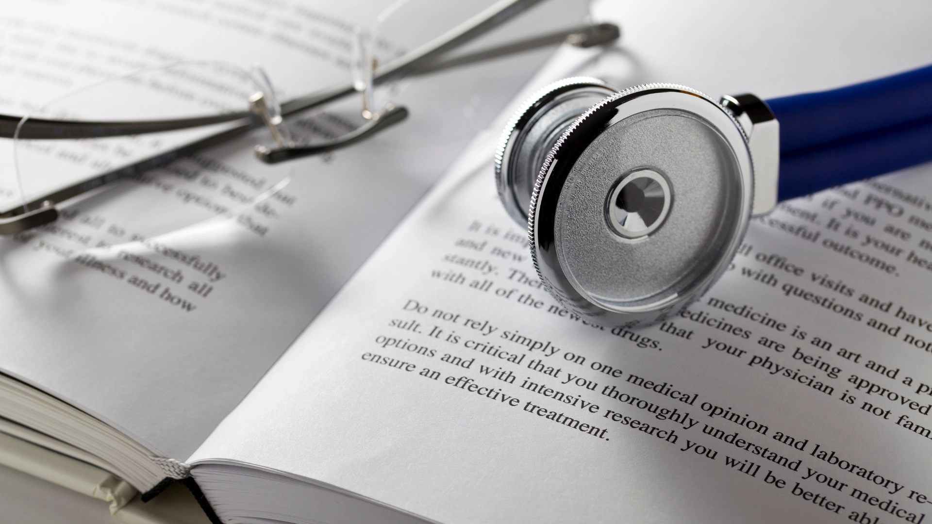 A stethoscope rests on top of an open book, with a blurred medical textbook visible in the background.