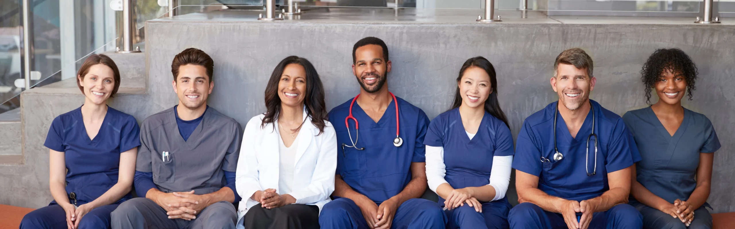 A group of seven healthcare professionals, including doctors and nurses wearing blue scrubs and one white doctor's jacket, are sitting and smiling together.