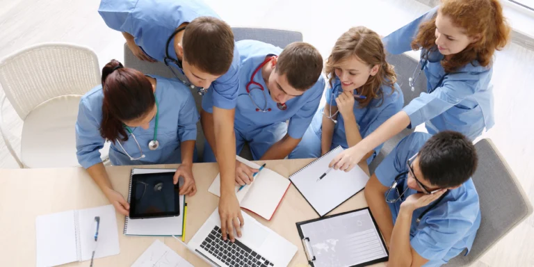 Overhead view of a diverse group of healthcare professionals wearing blue scrubs gathered around a table, collaborating over medical documents and a laptop.