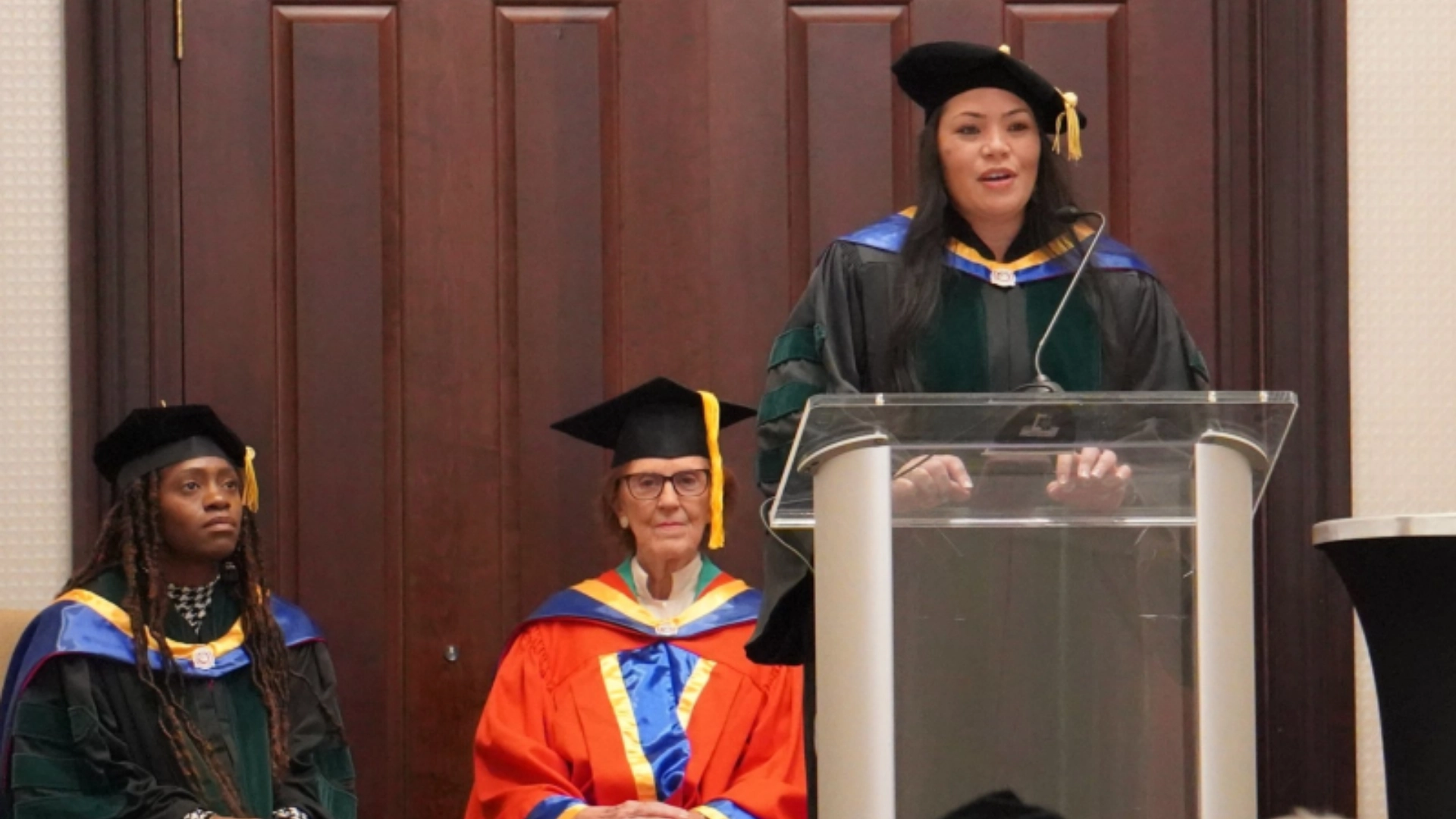 Graduation ceremony with a speaker in academic regalia addressing the audience at a podium, while two faculty members in caps and gowns sit behind her