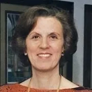 A close-up portrait of a smiling woman with light brown hair and fair skin. She has a gentle expression and is wearing an orange-reddish colored top.